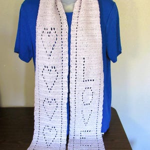 May include: A light pink crocheted scarf with the word "LOVE" and heart shapes incorporated into the design. The scarf is displayed on a mannequin and has a rectangular shape. The scarf is made of a soft, textured yarn.