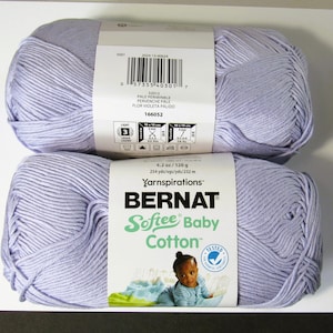 May include: Two skeins of pale periwinkle Bernat Softee Baby Cotton yarn stacked vertically. Each skein features a white paper label with a barcode, care instructions, and text indicating 4.2 oz, 120 g, and 254 yds, 232 m of light weight yarn.