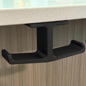 May include: Black metal hook that attaches under a shelf or desk. The hook has two prongs for hanging items.