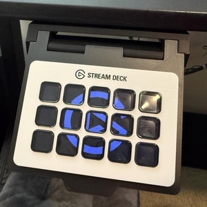 Puede incluir: Un Stream Deck blanco con 15 botones negros, cada uno con un icono azul. Los botones están dispuestos en una cuadrícula de 5x3. El Stream Deck está montado en un escritorio negro.
