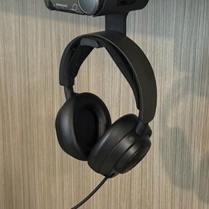 Angled view of a 3D printed black under-desk mount installed beneath a white desk, holding both the SteelSeries Arctis Nova Pro Wireless base station and the headset hanging from the integrated hook below. The setup showcases a clean, space-saving design with visible cable access and a secure fit.