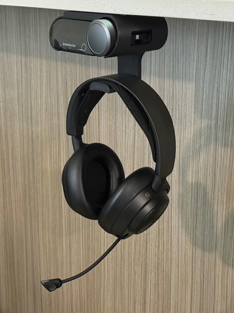 Angled view of a 3D printed black under-desk mount installed beneath a white desk, holding both the SteelSeries Arctis Nova Pro Wireless base station and the headset hanging from the integrated hook below. The setup showcases a clean, space-saving design with visible cable access and a secure fit.