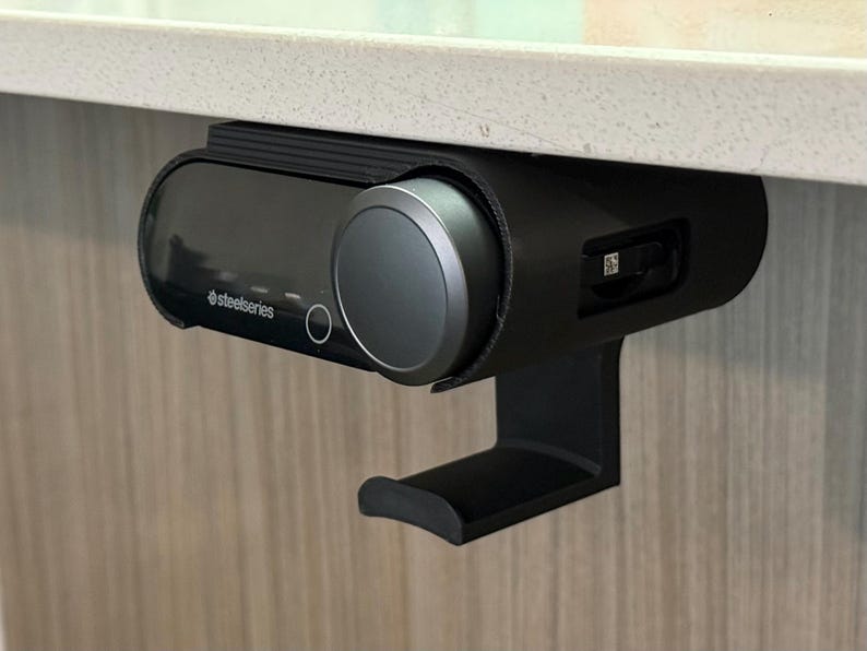 Black under-desk mount securely holding a SteelSeries Arctis Nova Pro Wireless base station, with a curved hook below designed to hang the headset. The 3D printed black mount is attached beneath a white desk surface, showcasing cable clearance and battery slot access.