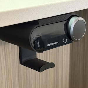 Close-up of a black under-desk mount holding a SteelSeries Arctis Nova Pro Wireless base station, with a built-in hook below for hanging the headset. The mount is 3D printed and neatly installed beneath a white desk surface, demonstrating a clean and space-saving setup.