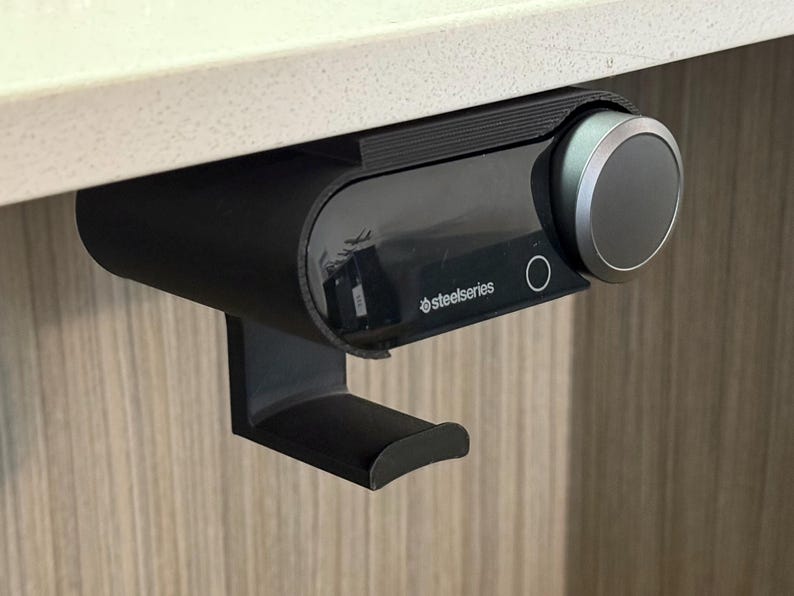 Close-up of a black under-desk mount holding a SteelSeries Arctis Nova Pro Wireless base station, with a built-in hook below for hanging the headset. The mount is 3D printed and neatly installed beneath a white desk surface, demonstrating a clean and space-saving setup.