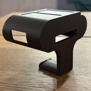 May include: A black 3D printed phone holder with a curved top and a rectangular base. The holder has two slots on the top for holding a phone.