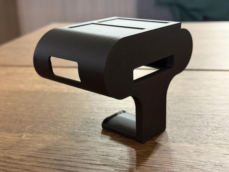 May include: A black 3D printed phone holder with a curved top and a rectangular base. The holder has two slots on the top for holding a phone.