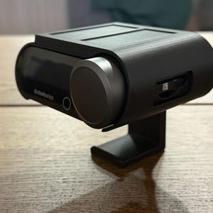 May include: A black, plastic case with a built-in stand for a small, black electronic device. The device has a small screen and a round button on the side. The case has a slot for a memory card.