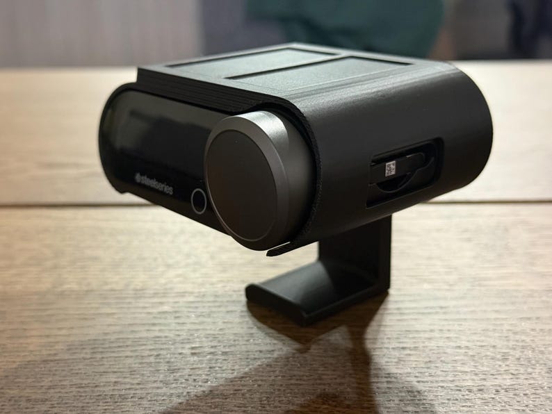 May include: A black, plastic case with a built-in stand for a small, black electronic device. The device has a small screen and a round button on the side. The case has a slot for a memory card.