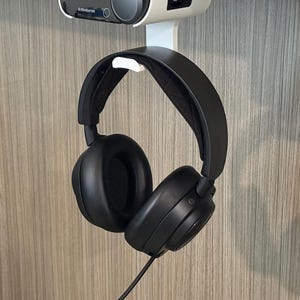 Angled view of a 3D printed white under-desk mount installed beneath a white desk, holding both the SteelSeries Arctis Nova Pro Wireless base station and the headset hanging from the integrated hook below. The setup showcases a clean, space-saving design with visible cable access and a secure fit.