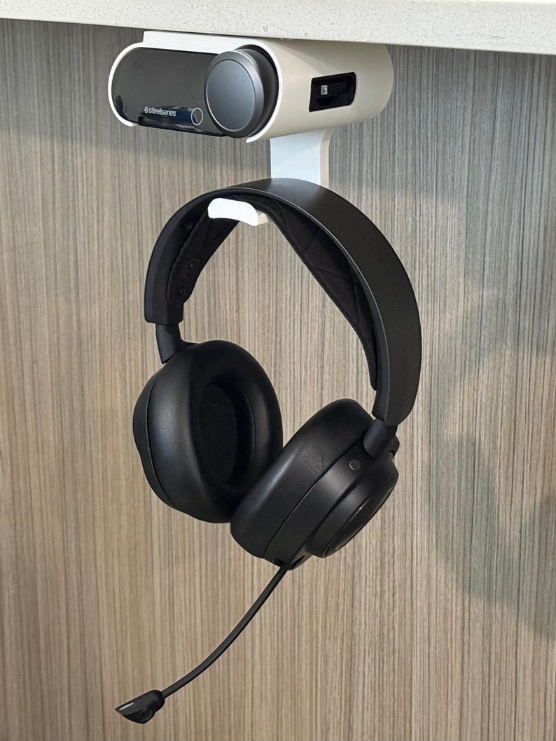 Angled view of a 3D printed white under-desk mount installed beneath a white desk, holding both the SteelSeries Arctis Nova Pro Wireless base station and the headset hanging from the integrated hook below. The setup showcases a clean, space-saving design with visible cable access and a secure fit.