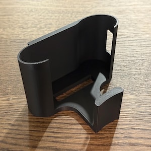 May include: A black plastic phone holder with a curved design. The holder has a slot for a phone and a stand on the bottom.