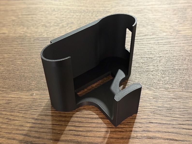 May include: A black plastic phone holder with a curved design. The holder has a slot for a phone and a stand on the bottom.