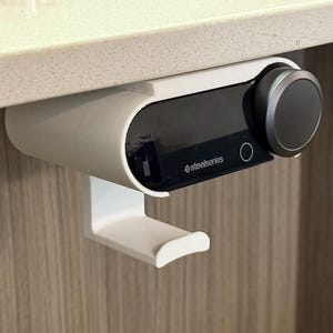 Close-up of a white under-desk mount holding a SteelSeries Arctis Nova Pro Wireless base station, with a built-in hook below for hanging the headset. The mount is 3D printed and neatly installed beneath a white desk surface, demonstrating a clean and space-saving setup.