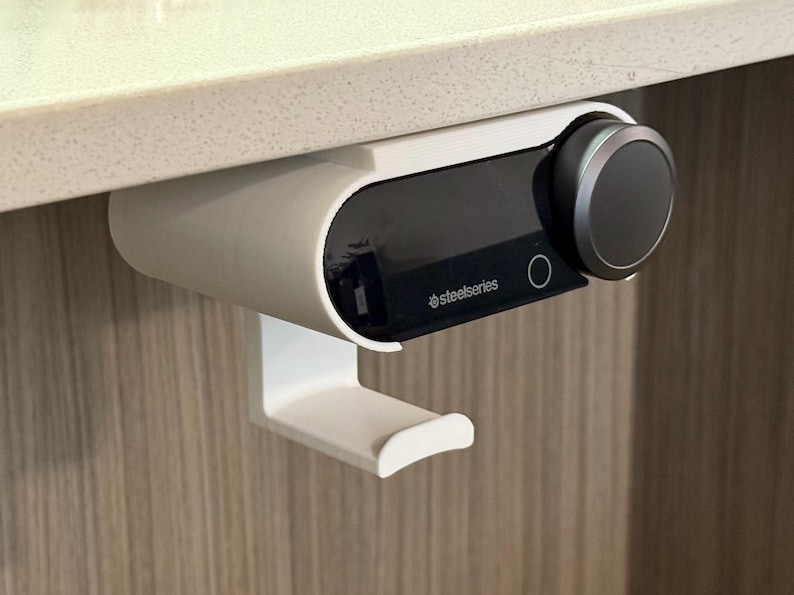 Close-up of a white under-desk mount holding a SteelSeries Arctis Nova Pro Wireless base station, with a built-in hook below for hanging the headset. The mount is 3D printed and neatly installed beneath a white desk surface, demonstrating a clean and space-saving setup.