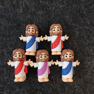 May include: Five small figurines of Jesus with outstretched arms. Each figure has brown hair and a beard, wearing a white robe with a colored sash in blue, red, or purple. The figurines are on a dark gray surface.