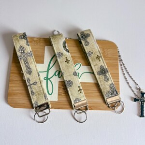 May include: Three beige keychains with ornate cross patterns and silver hardware are displayed on a wooden board with the word "faith" written in green script. A silver necklace with a cross pendant is also visible.