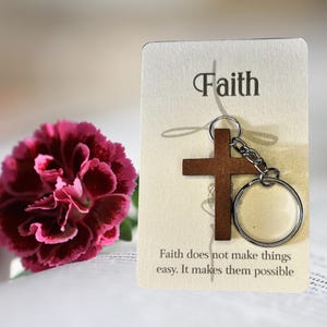 Könnte beinhalten: Ein Schlüsselanhänger aus Holz in Form eines Kreuzes mit einem silbernen Ring und einer Kette. Der Schlüsselanhänger ist an einer Karte mit dem Wort "Glaube" und dem Zitat "Glaube macht die Dinge nicht einfach. Er macht sie möglich." befestigt.