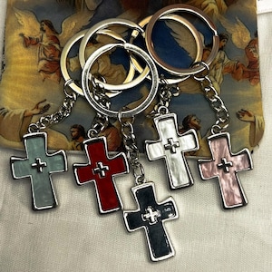 以下が含まれることがあります： 6つの十字架型のキーホルダーのコレクション。各十字架は、水色、赤、白、黒、ピンクと異なる色です。十字架はチェーンで銀色のキーリングに取り付けられています。背景は天使のデザインの生地です。