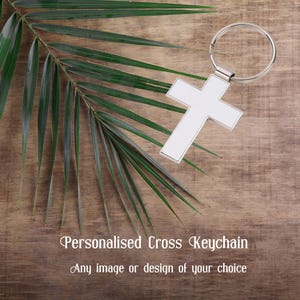 Puede incluir: Un llavero de metal blanco en forma de cruz con un anillo plateado. La cruz está sobre una superficie de madera con una hoja de palma verde.