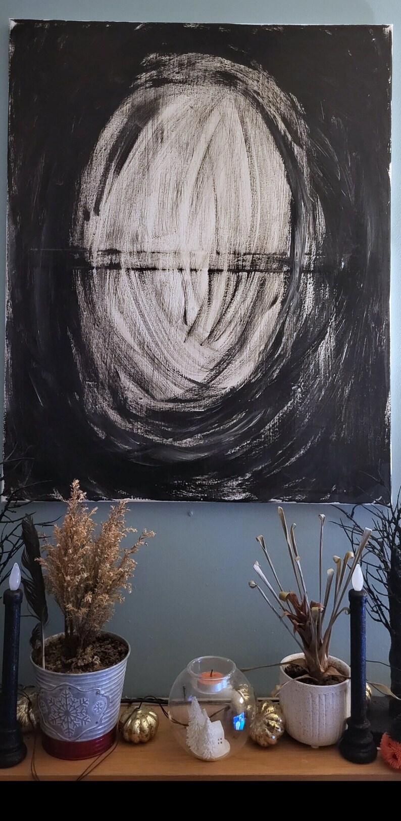 Op de afbeelding: Een abstract zwart-wit schilderij met een cirkelvorm in het midden. Het schilderij hangt aan een muur met een lichtblauwe achtergrond. Op de voorgrond staan planten en kaarsen.