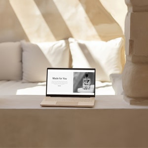 May include: A beige laptop displaying a website with the text "Made for You" and a black and white image of a perfume bottle. The laptop is on a white surface, with white pillows in the background. Sunlight streams in from the window.