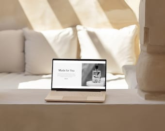 May include: A beige laptop displaying a website with the text "Made for You" and a black and white image of a perfume bottle. The laptop is on a white surface, with white pillows in the background. Sunlight streams in from the window.