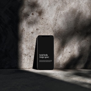 Op de afbeelding: Een zwarte smartphone met een leeg scherm leunt tegen een getextureerde betonnen muur. De telefoon toont de tekst "Social Media Design Agency" in het wit. De scène wordt verlicht met schaduwen, wat een minimalistische esthetiek creëert.