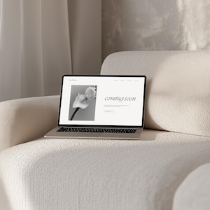 May include: A laptop computer on a white couch with a website open that says "coming soon" and a black and white photo of a flower.