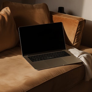 May include: A silver laptop computer with a black screen is open and resting on a brown couch. The couch is covered in a soft, brown fabric and has a pillow behind the laptop.