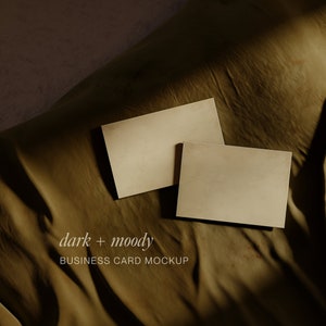 May include: Two blank business cards on a brown fabric background. The text "dark + moody BUSINESS CARD MOCKUP" is printed below the cards.