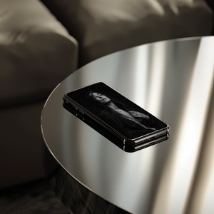 以下が含まれることがあります： 画面に白黒の人物の写真が表示された黒いスマートフォン。スマートフォンは反射面の上に置かれています