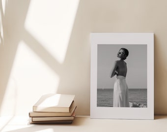 May include: A black and white photograph of a person standing on a beach, wearing a white skirt and looking over their shoulder. The photo is framed in a white frame and is sitting on a shelf.
