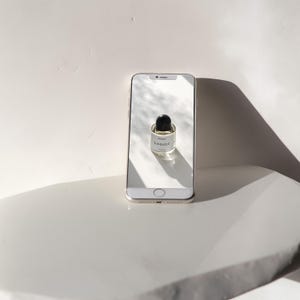 May include: A white smartphone with a black screen displaying a small glass bottle of perfume with a black cap. The bottle is labeled "SUNDAZED" in black text.