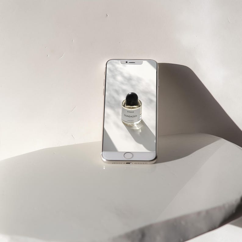 May include: A white smartphone with a black screen displaying a small glass bottle of perfume with a black cap. The bottle is labeled "SUNDAZED" in black text.
