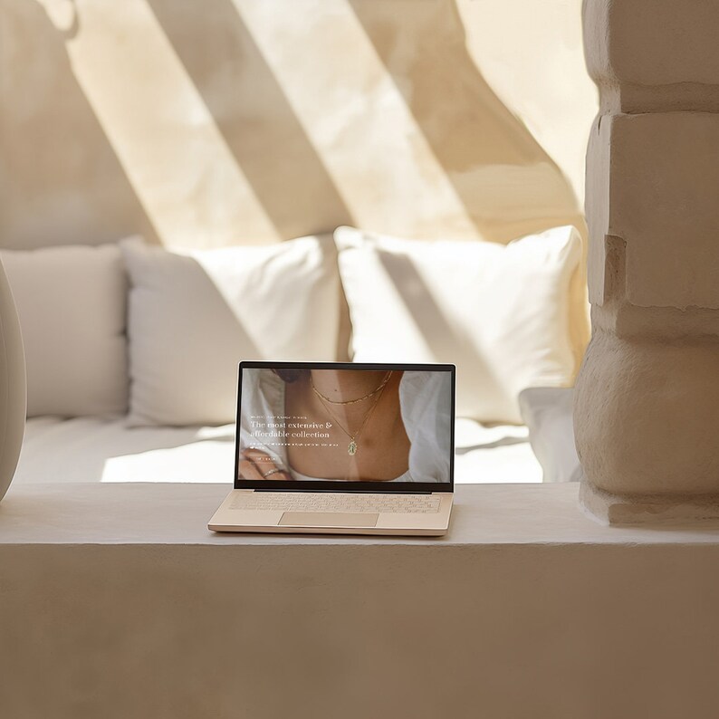 May include: A beige laptop displaying a website with jewelry. The screen shows a woman wearing necklaces. The text on the screen reads, "The most extensive & affordable collection." The laptop is open on a white ledge, with white pillows in the background.