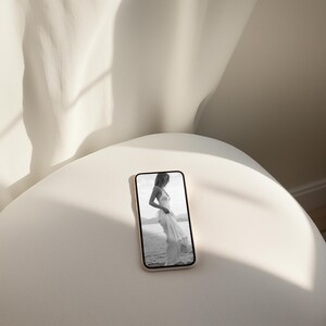 May include: A black and white photo of a person wearing a white dress, displayed on a smartphone screen. The phone is sitting on a white surface.