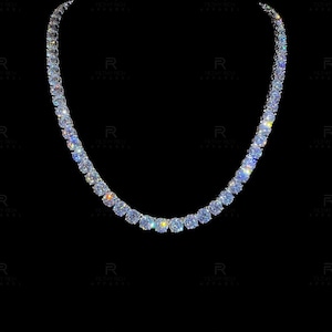 May include: A tennis necklace featuring a continuous row of round, brilliant-cut clear stones set in a metallic frame. The stones exhibit high clarity and light reflection, creating a sparkling effect against the dark background of the image.