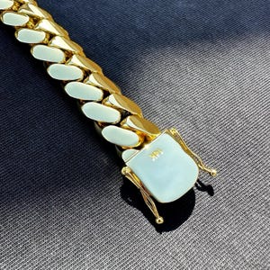 14K gold-plated Miami Cuban link bracelet with box clasp, polished finish, and secure side locks, displayed on dark fabric under natural sunlight