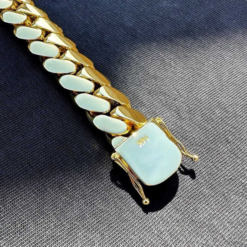14K gold-plated Miami Cuban link bracelet with box clasp, polished finish, and secure side locks, displayed on dark fabric under natural sunlight