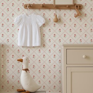 Könnte beinhalten: Ein Kinderzimmer mit einem weißen Kleid, das an einer Holzstange hängt, einer Stoffgans und einem grauen Koffer. Die Tapete hat ein Blumenmuster mit roten Blumen und rosa Gitterlinien. Eine beige Kommode und ein Kinderbett sind ebenfalls zu sehen.