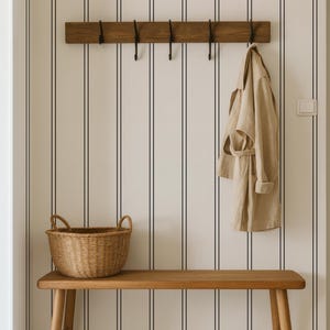 A wooden bench with a wicker basket sits against a striped wall, with a coat hanging on a wooden coat rack above it.
