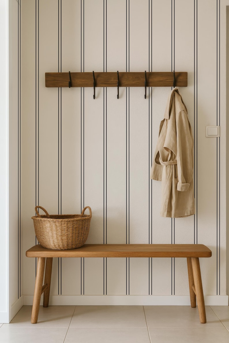 A wooden bench with a wicker basket sits against a striped wall, with a coat hanging on a wooden coat rack above it.