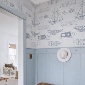 a room with a wooden bench, a white hat on a hook, and a wall with a pattern of sailboats and other nautical illustrations. The room appears to be a living or dining area, with a couch and a coffee table visible in the background.