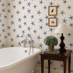 Pode incluir: Uma casa de banho com uma banheira branca com pés de garra, uma mesa lateral de madeira escura e uma parede coberta por um padrão de estrelas do mar azuis sobre um fundo creme. Uma lâmpada e quadros emoldurados estão sobre a mesa. A palavra "INTERIORS" é visível num livro.