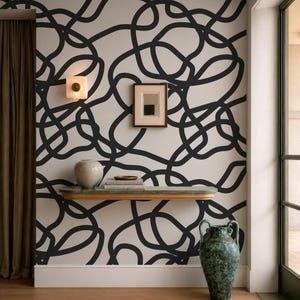 May include: Wallpaper featuring a continuous black line pattern on a light cream background. A floating shelf holds a ceramic vase and books, while a framed print hangs above. A large, weathered green urn sits on the floor to the right.
