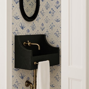 May include: A black wall-mounted sink with a brass faucet and plumbing, and a white towel. An oval black mirror hangs above the sink. The wall is covered in a floral wallpaper with blue and white flowers.