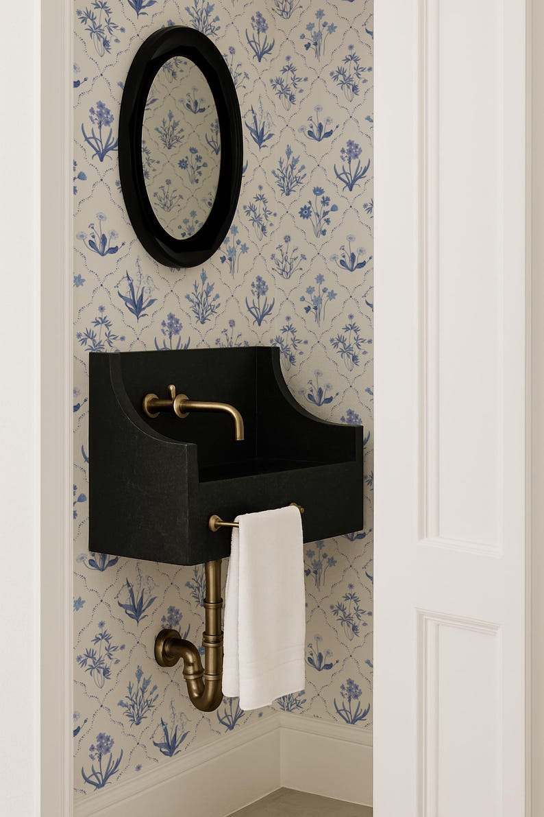 May include: A black wall-mounted sink with a brass faucet and plumbing, and a white towel. An oval black mirror hangs above the sink. The wall is covered in a floral wallpaper with blue and white flowers.