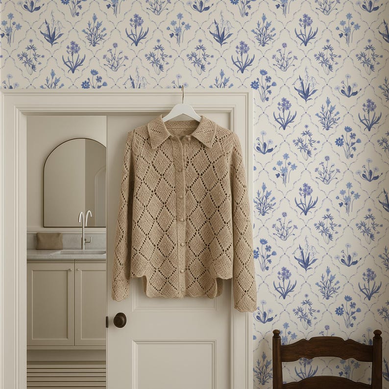 May include: A beige, open-weave cardigan with a scalloped hem hangs on a white hanger. The cardigan has a diamond pattern and is buttoned up. The background features a floral wallpaper in white and blue, and a glimpse of a bathroom.