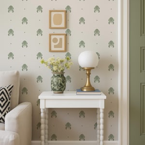 Könnte beinhalten: Innenraum mit einem weißen Beistelltisch mit Lampe, Vase und Büchern. Die Wand ist mit einem sich wiederholenden grünen Blumenmuster verziert. Zwei gerahmte Kunstwerke hängen über dem Tisch. Ein weißes Sofa mit gemustertem Kissen ist zu sehen.